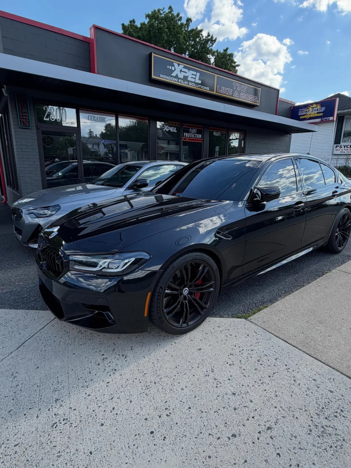 Black BMW M5 with tinted windows in front of the XPEL Window Film Solutions storefront